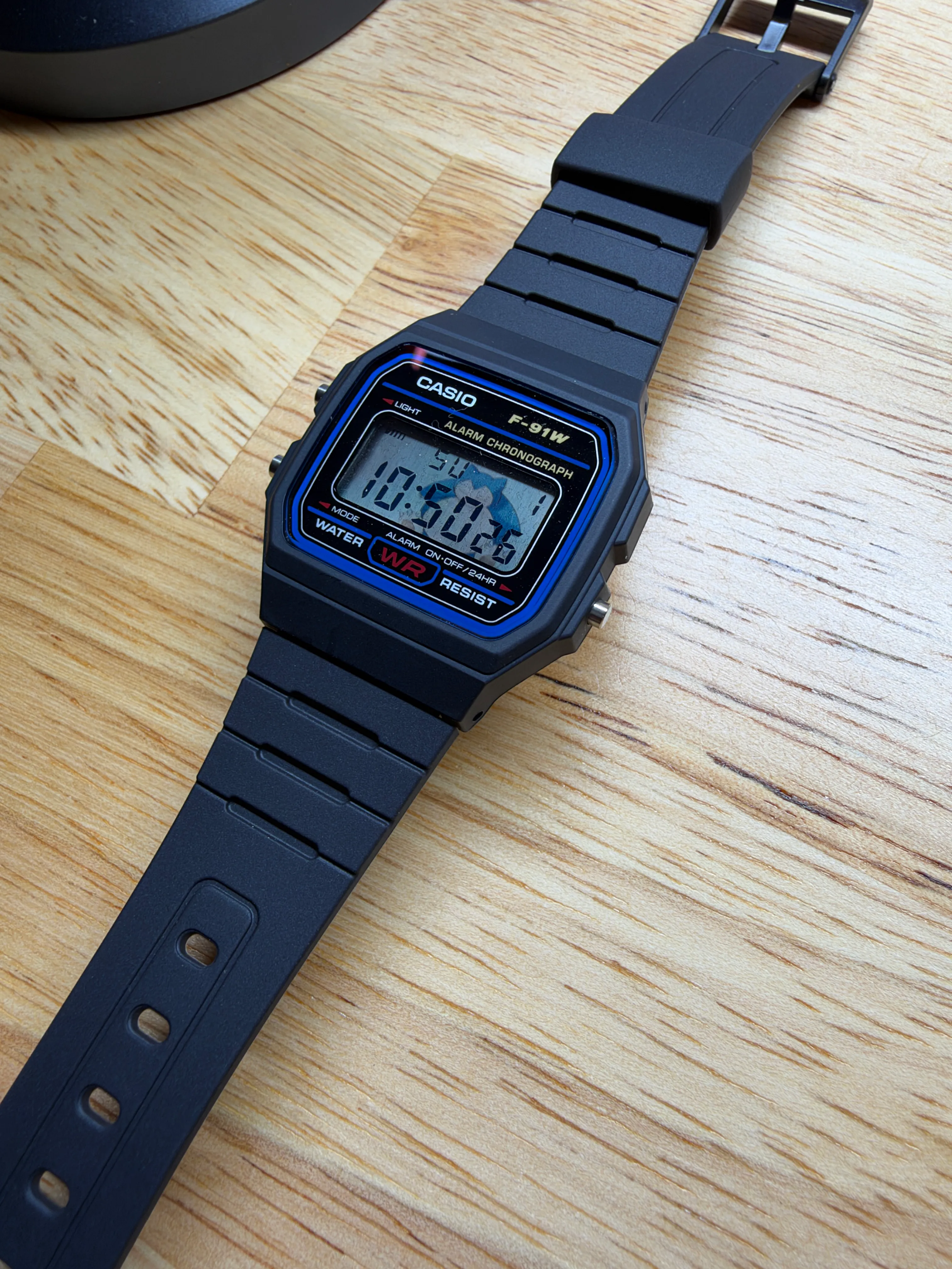 Angled close-up of the black Casio F-91W with Snorlax artwork visible behind the LCD, resting on a light wood surface