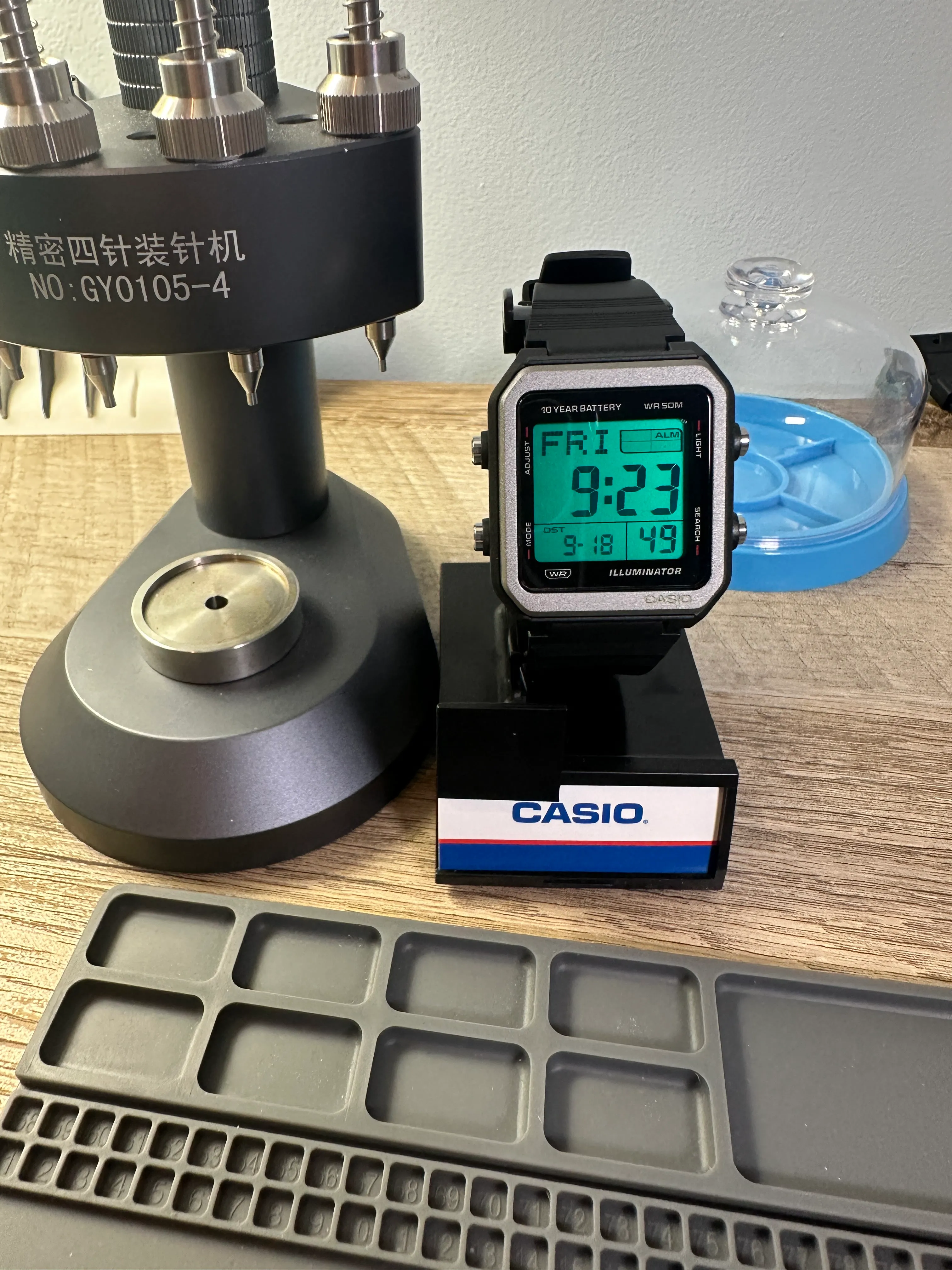 A Casio W-221H with a black case and silver bezel on a Casio stand, the display glowing green, beside a hand-setting press